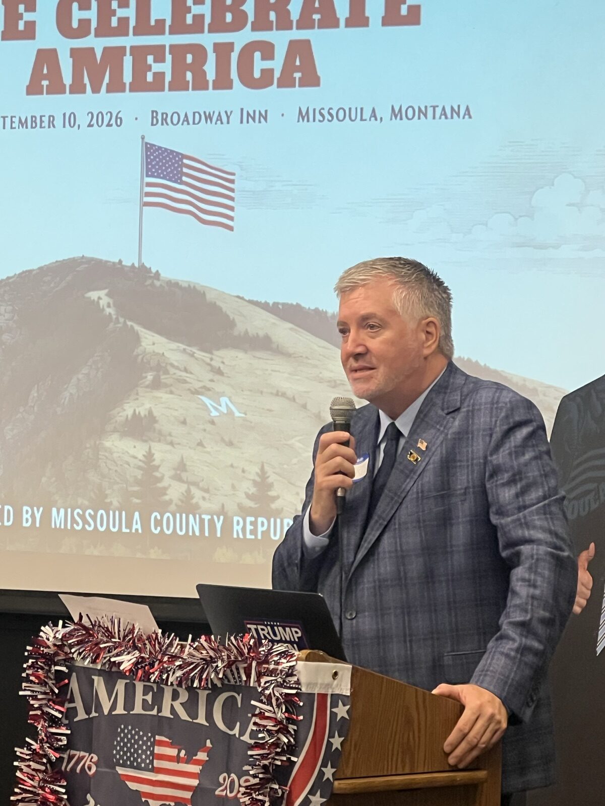 Chris Warden emcees Celebrate America at the podium, with the event backdrop behind him.