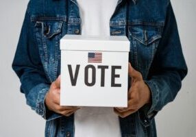Person in denim holding a ballot box with a US flag, emphasizing voting rights.